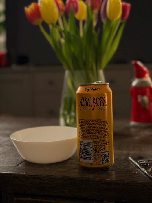 Burk med Albatross NEIPA 7.0% på ett bord, bredvid en vit skål och en fjärrkontroll. Bakgrund med tulpaner och en röd prydnad.