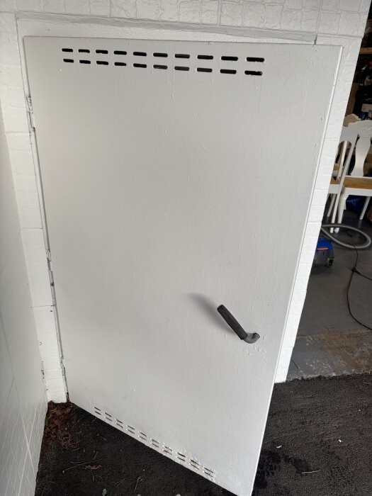 En vit dörr med ventilationshål och en enkel dörrhandtag, installerad nära ett mörkt betonggolv.