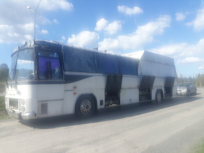 En gammal vit buss med flera öppna sidopaneler står parkerad på en landsväg under en blå himmel med moln.