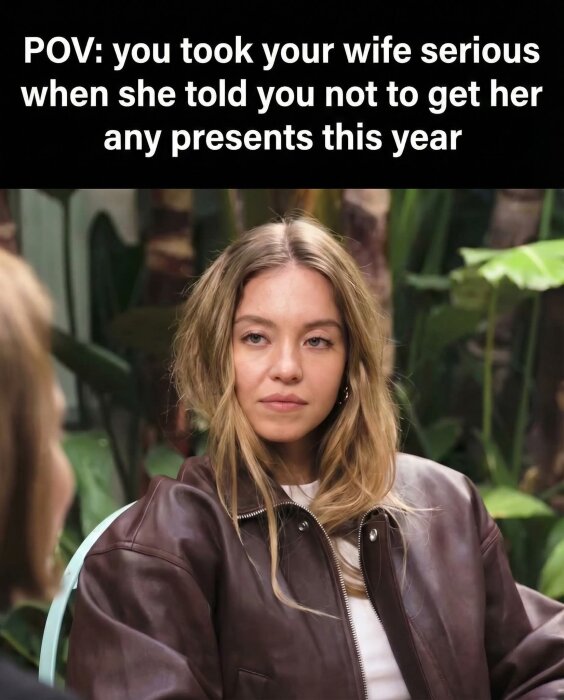 Kvinna i brun jacka i en trädgårdsmiljö ser seriös ut, med bildtexten "POV: you took your wife serious when she told you not to get her any presents this year".