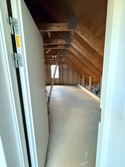 Vind i ett trähus under renovering, med synliga bjälkar och ett öppet fönster i slutet av rummet.