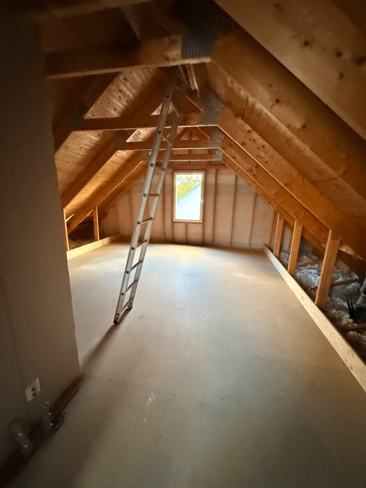 Vind med träbjälkar, spånskivegolv och stege, sedd ovanifrån. Ett litet fönster ger ljusinsläpp. Utförs för ombyggnad till hemmabio.