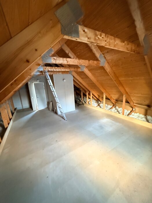 Vind med snedtak, träbjälkar och ett golv av spånskivor under renovering, förvandlas till hemmabiorum i ett trähus. Stege och isolering synliga.