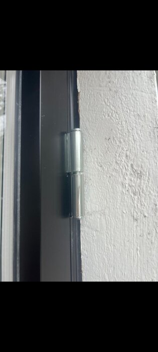 Metallgångjärn monterat på en svart dörrkarm intill en vit vägg med textur.