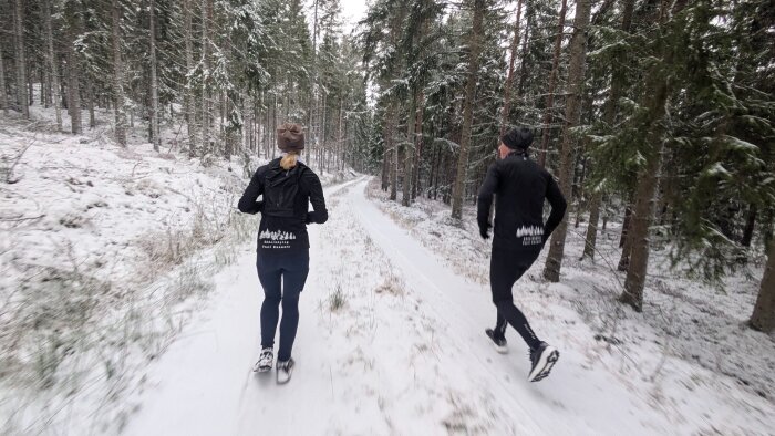 Två personer joggar på en snötäckt skogsstig omgivna av höga träd.