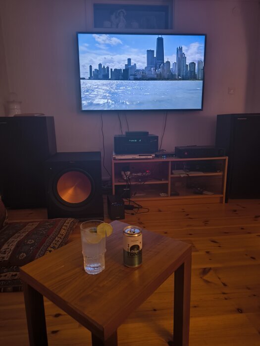 Hemmbio med stor TV, högtalare och subwoofer på trägolv; drink och tonicburk på bord framför.