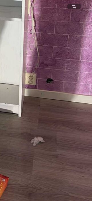 Lila tegelvägg med ett eluttag och kablar, hål i väggen. Trägolv med pappersbitar och en vit hylla till vänster.