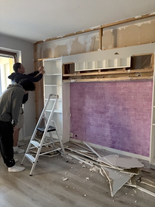 Två personer renoverar ett rum med rosa tegelvägg genom att riva en vägg med hjälp av en stege, omgiven av bråte på golvet.