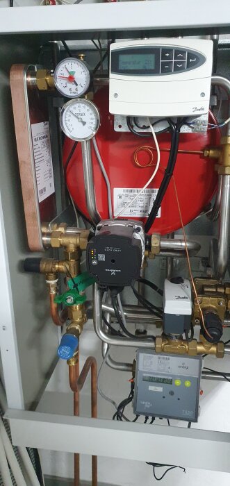 Fjärrvärmecentral med mätinstrument, rör och en grön kran med etiketten "påfyllning vatten.