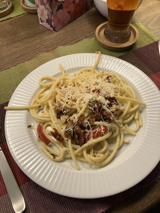 Tallrik med pasta toppad med riven ost, soltorkade tomater och kryddor, på ett dukat bord bredvid ett glas öl.