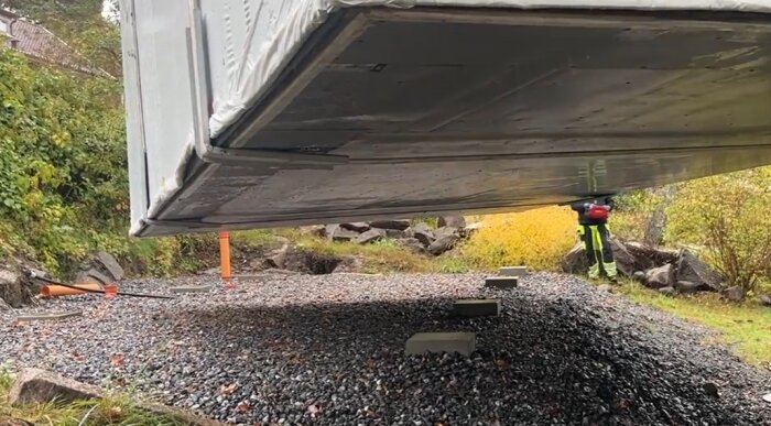 En byggnad placeras på markstenar ovanför en grusad yta i en trädgårdsmiljö, med en person som inspekterar under.