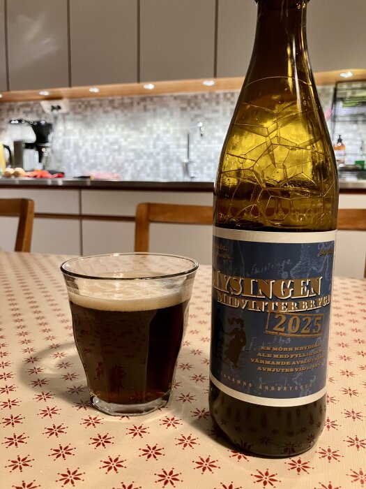 Flaska och glas med mörk öl på ett bord i kök med mönstrad duk, bakgrund med skåp och köksutrustning.