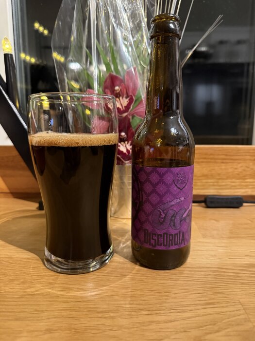 Flaska och glas med mörk ölsort Discordia på ett bord, blomma i bakgrunden.
