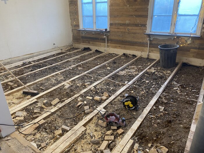 Rum under renovering med upplagda golvbjälkar, verktyg och materialskräp på marken. Väggar halvklädda och två fönster synliga.