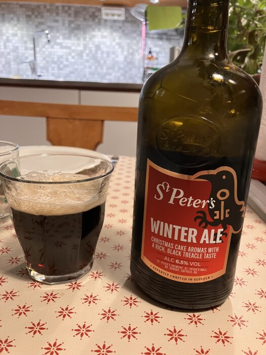 Flaska med St Peter's Winter Ale bredvid ett glas mörk öl på ett bord med rött och vitt mönstrat duk. Bakgrund av köksmiljö.
