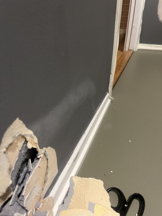 Skadat område i grå vägg nära golvet, där färg och gips har lossnat och blottar underliggande material. En svart sax ligger på golvet bredvid.