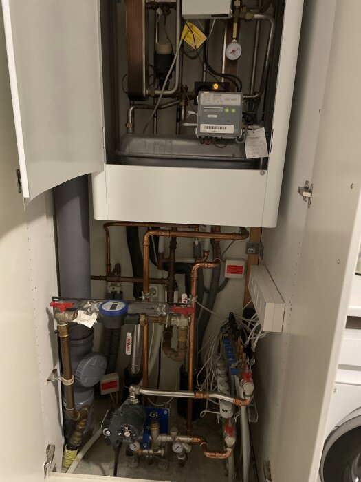 Värmepumpsinstallation med rörsystem och mätare i ett skåp med öppna dörrar.