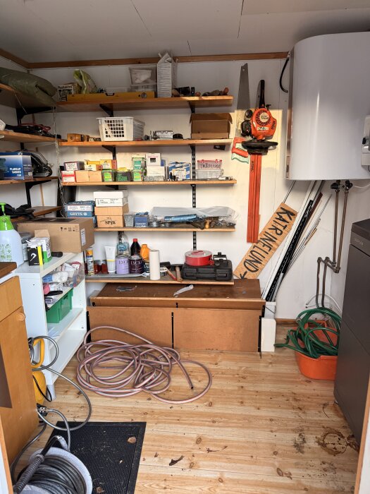 Fristående förråd med hyllor fyllda med verktyg och material. Varmvattenberedare till höger och hoprullade slangar på träplankgolv.