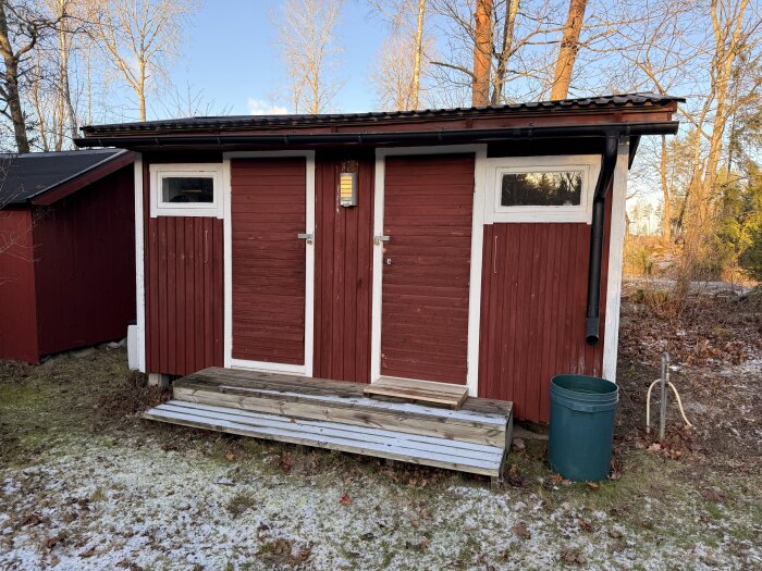 Rött fristående förråd med två dörrar, vita karmar och kringliggande träd. Grön tunna bredvid, vinterlandskap. Möjligt projekt för bastuombyggnad.