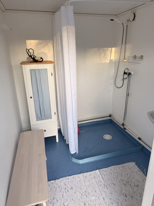 En duschkabin med draperi i ett rum med blått golv, vit vägg och ett litet, vitt skåp med trådkorg på väggen.