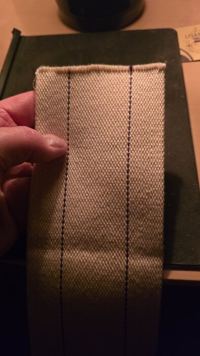 En hand håller ett stycke beige vävt tyg med två vertikala svarta linjer, mot en suddig bakgrund av ett skrivbord och en anteckningsbok.