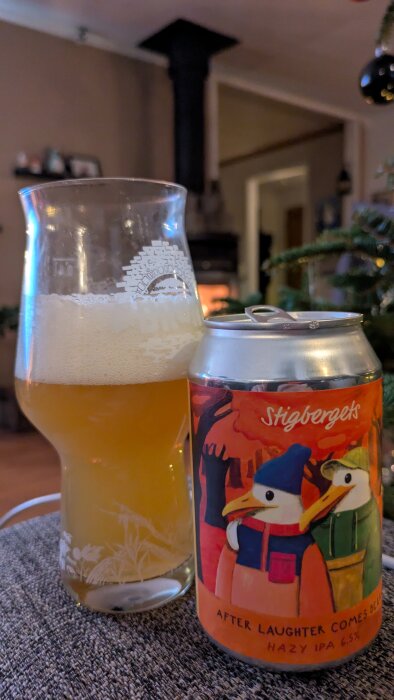 Ett glas med hazy IPA och en ölburk från Stigbergets med färgglad etikett, i bakgrunden en eldstad och julgran.