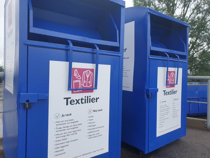 Två blå containrar för textilåtervinning med instruktioner för vad som får kastas, fotograferade utomhus en fuktig dag.