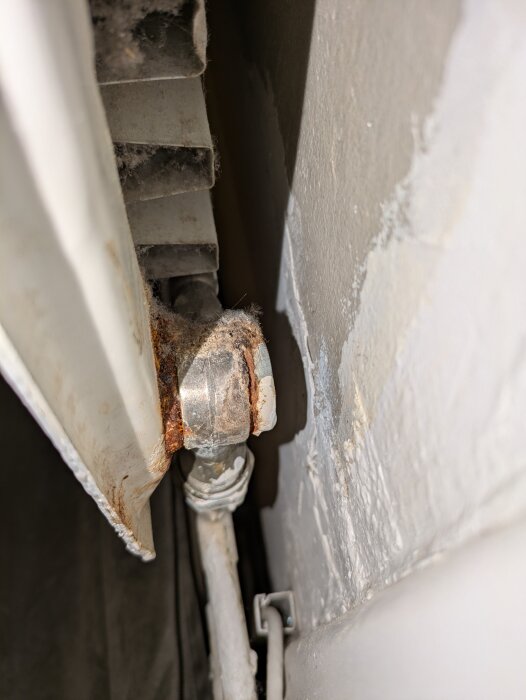 Rostigt röranslutning på gammal radiator med smuts och damm, rör löper längs väggen i gammalt hus, fokus på underhåll behövs.