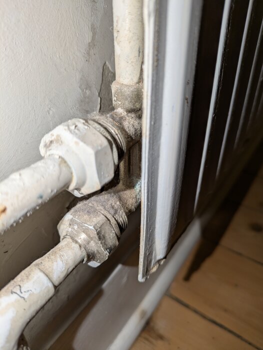 Röranslutning under radiator, del av värmesystem. Koppel och ventil på rör synliga, rör går in i väggen. Golv av trä. Lätt slitage syns.