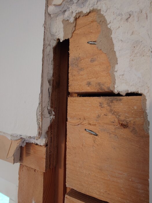 Detalj av en husvägg med borttagen gips som avslöjar äldre träpanel och isolering. Skador och bristande plast finns synligt.