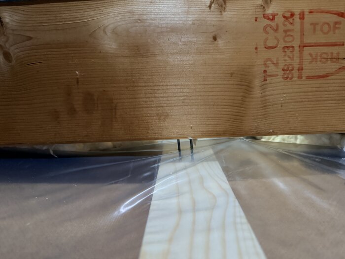 Bilden visar en närbild av en träregel med inskriptionen "C24" och plastfolie ovanpå en planka, sannolikt i ett byggprojekt.