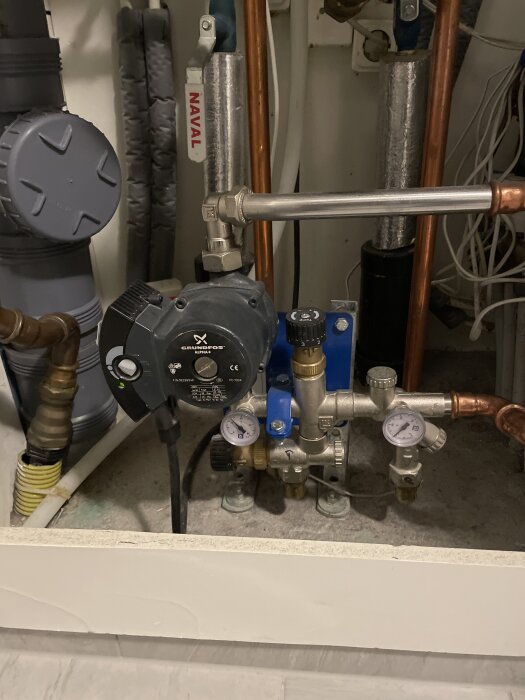 En rörinstallation med pumputrustning och rörkopplingar i ett hem, troligen del av ett värmesystem med fokus på en Grundfos-pump.