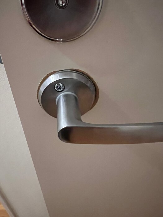 Dörrhandtag i borstad krom med otymplig monteringsbricka, synlig kant där täckbrist på tidigare större cylinder orsakat problem.