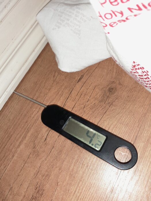 Temperaturdisplay visar 4,2°C på termometer placerad på trägolv i hörn, indikerar kallt hus.