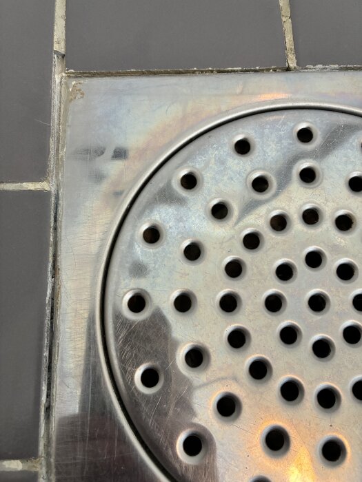 Nära bild av en golvbrunn i metall med omgivande mörka klinkerplattor, där fogarna runt brunnen är i fokus.