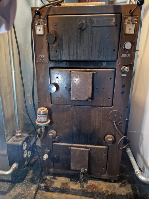 Gammal panna i källare med rostig yta, tillhörande rör och instrumentpanel med mätare och rattar.