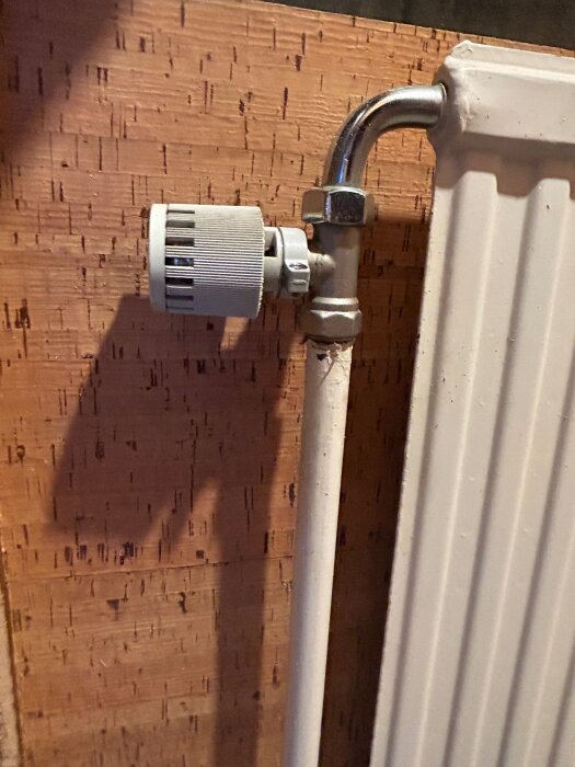 Bilden visar ett vitt element anslutet till en vägg med träliknande panel, med ett termostatvred på pipeline.