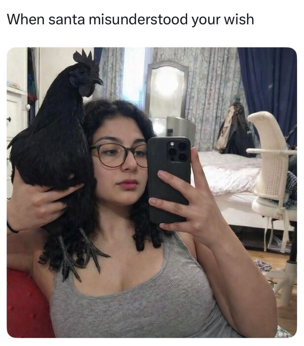 Person tar en selfie i en spegel och håller en svart höna vid huvudet i ett sovrum med säng och stol i bakgrunden.