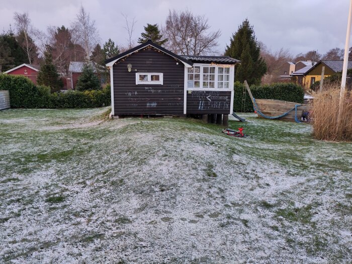 Litet hus på gård med lätt snötäckt gräsmatta, omgiven av buskar och träd.