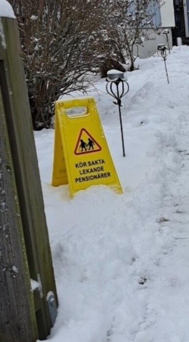 Gult varningsskylt i snöig trädgård med texten "Kör sakta lekande pensionärer", buskar och gång i bakgrunden.