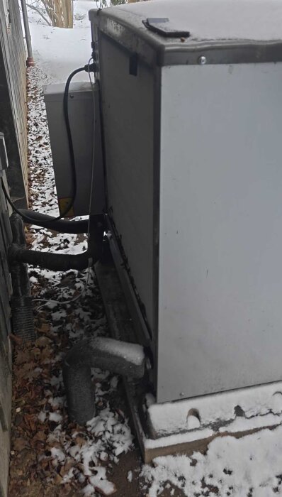 Värmepumpens utedel placerad på snötäckt mark på norrsidan av ett hus med stora isolerade rör anslutna.