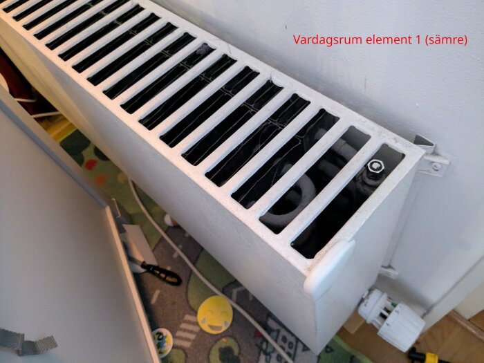 Vardagsrumselement med ventil och termostat mot vit vägg, delvis dolt lock, visas ovanifrån i närbild.