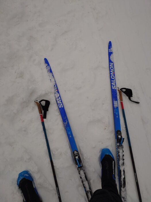 Blå längdskidor och stavar på snötäckt mark med blå skidpjäxor närbild.