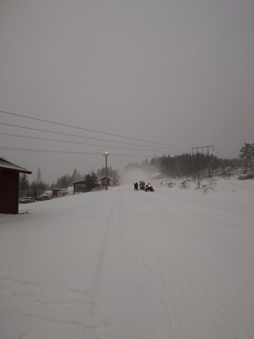 Snöig väg med traktor och person bredvid, omgivna av vinterlandskap och röda byggnader i bakgrunden, under en gråmulen himmel.