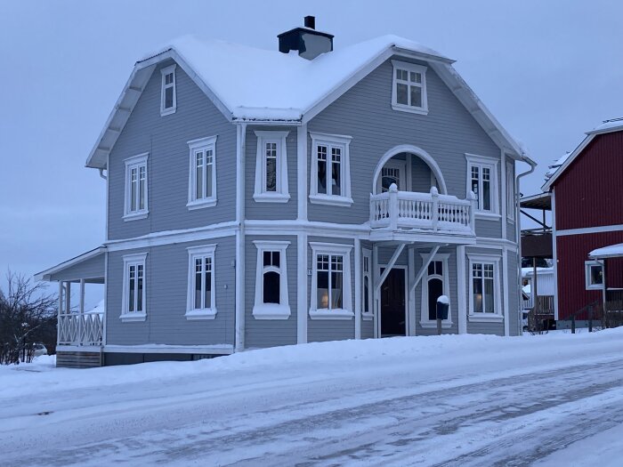Ett grått 20-talshus med snö på tak och mark, vit träbalkong och detaljer. Husets utbyggnad syns tydligt.