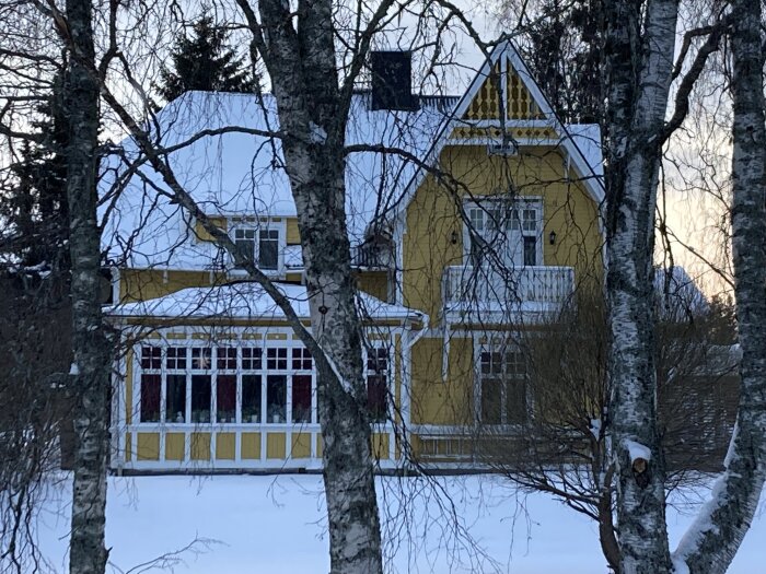Gult 20-talshus med snötäckt tak och glasad utbyggnad, omgivet av träd i vinterlandskap.