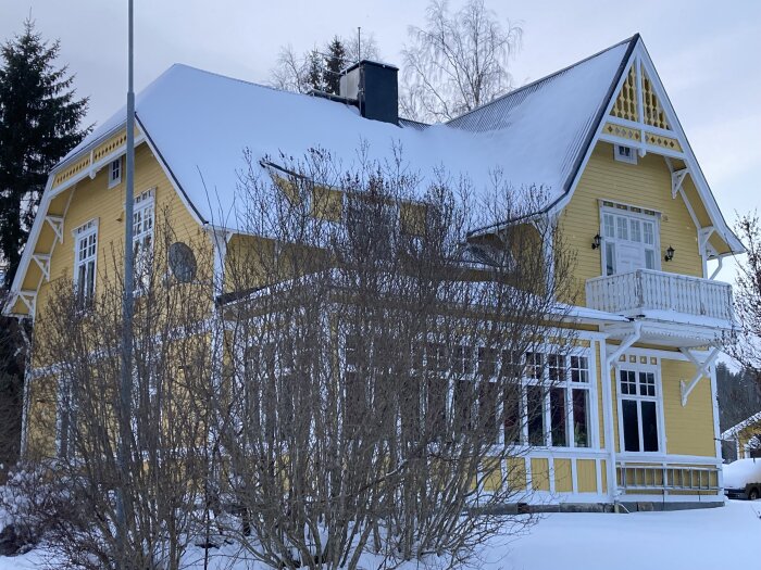 Gult 20-talshus med snötäckt tak och veranda, omgivet av vinterträd och buskar.