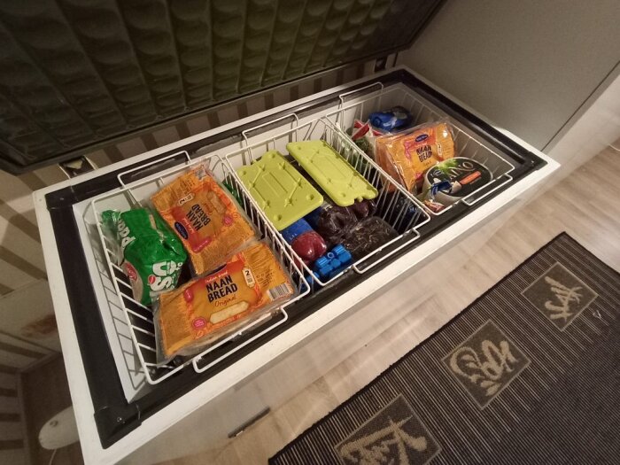 Öppen frysbox med sorterade matvaror i korgar, inklusive naanbröd, fryspåsar och plastbehållare, på ett ljust trägolv.