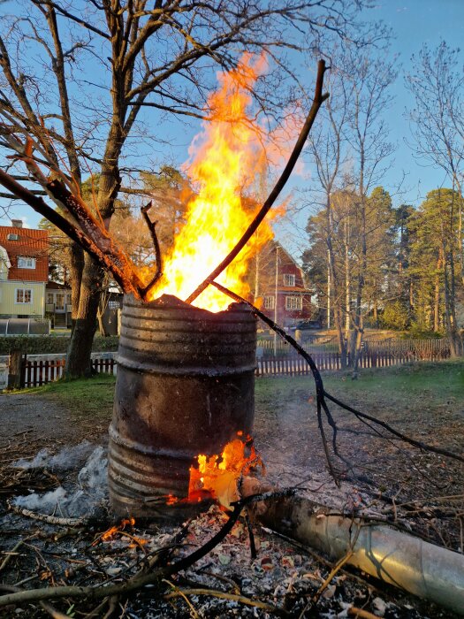Eld i en stor tunna på tomten med kvistar och sly, höga lågor mot en bakgrund av träd och hus i dagsljus.