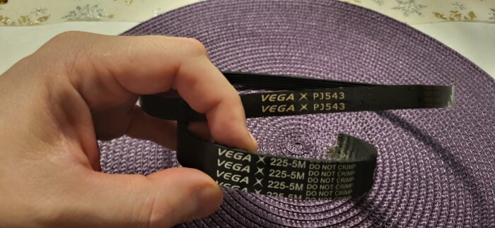 Hand som håller en svart tandrem från Vega med texten "PJ543" och "DO NOT CRIMP" på ett lila underlag.